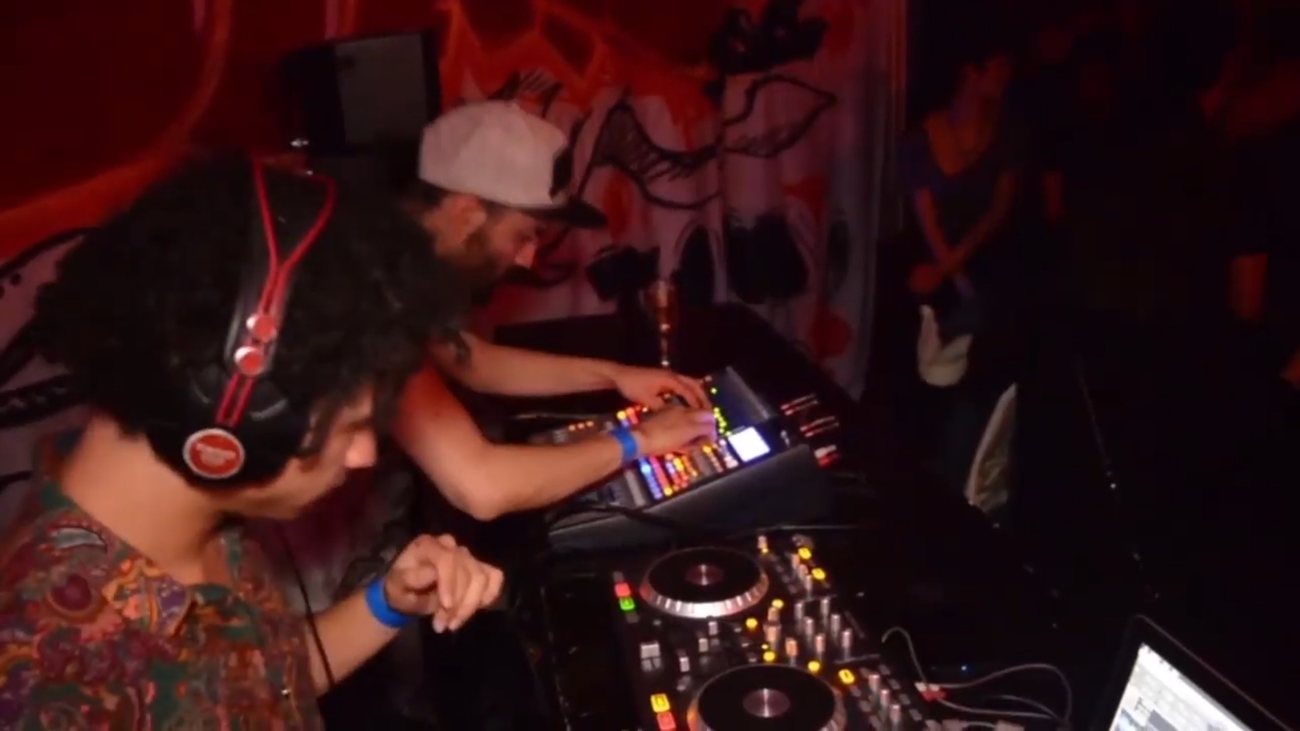 Two DJs at the decks in a red-lit booth with painted backdrop