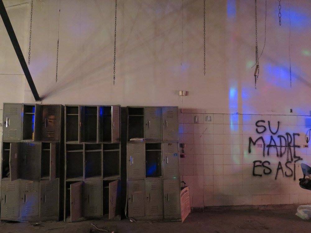 Row of old lockers under blue light, hanging chains, graffiti wall reading 'su madre es así'