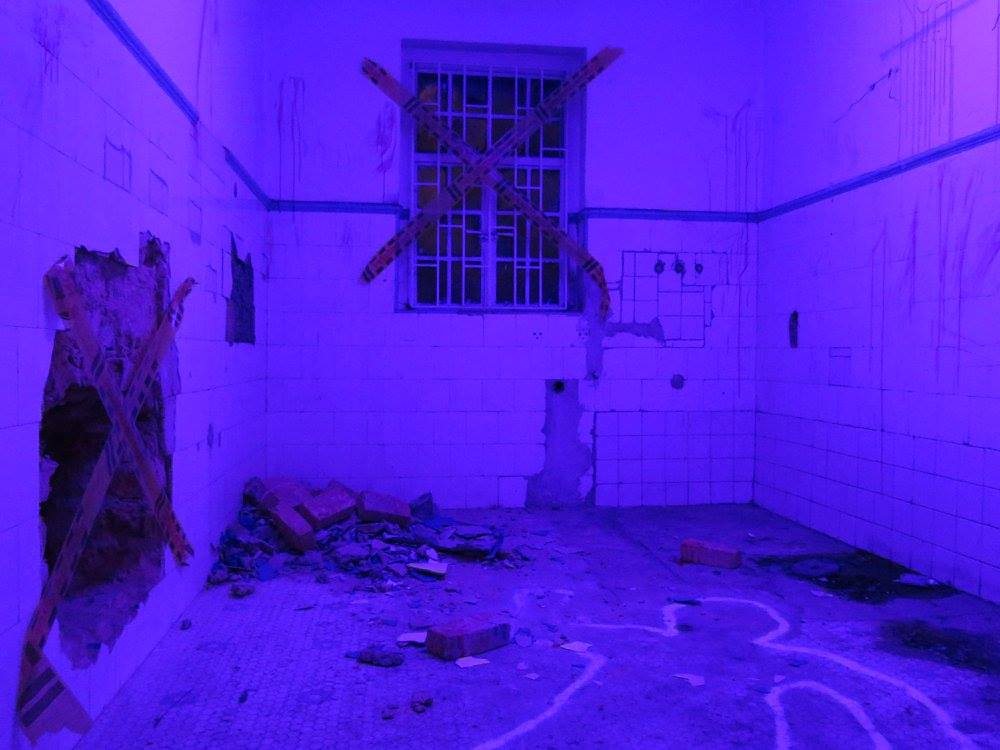 Tiled room bathed in purple light, barred window crossed with tape, rubble on the floor