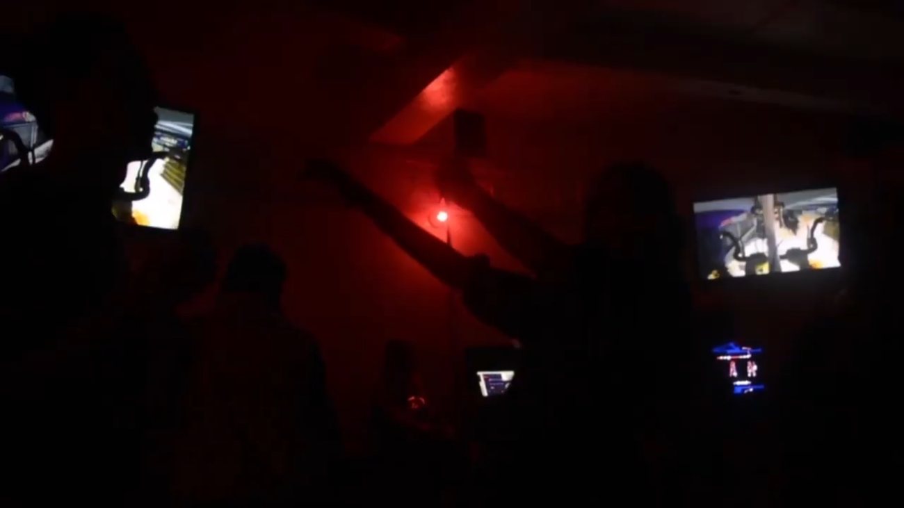 Dark room lit red, screens on the walls, silhouettes with arms raised