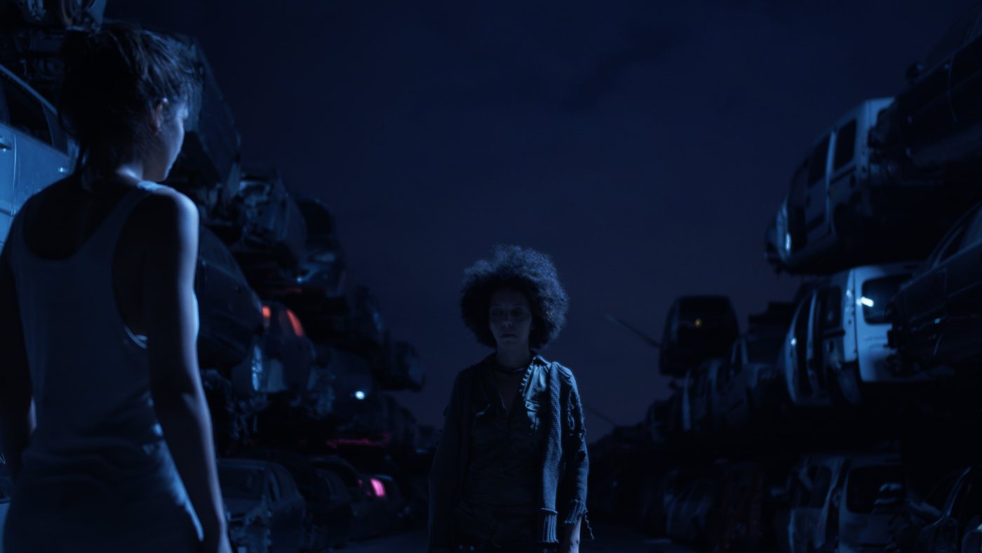 Ohnmacht — two women face each other in a blue-lit junkyard at night