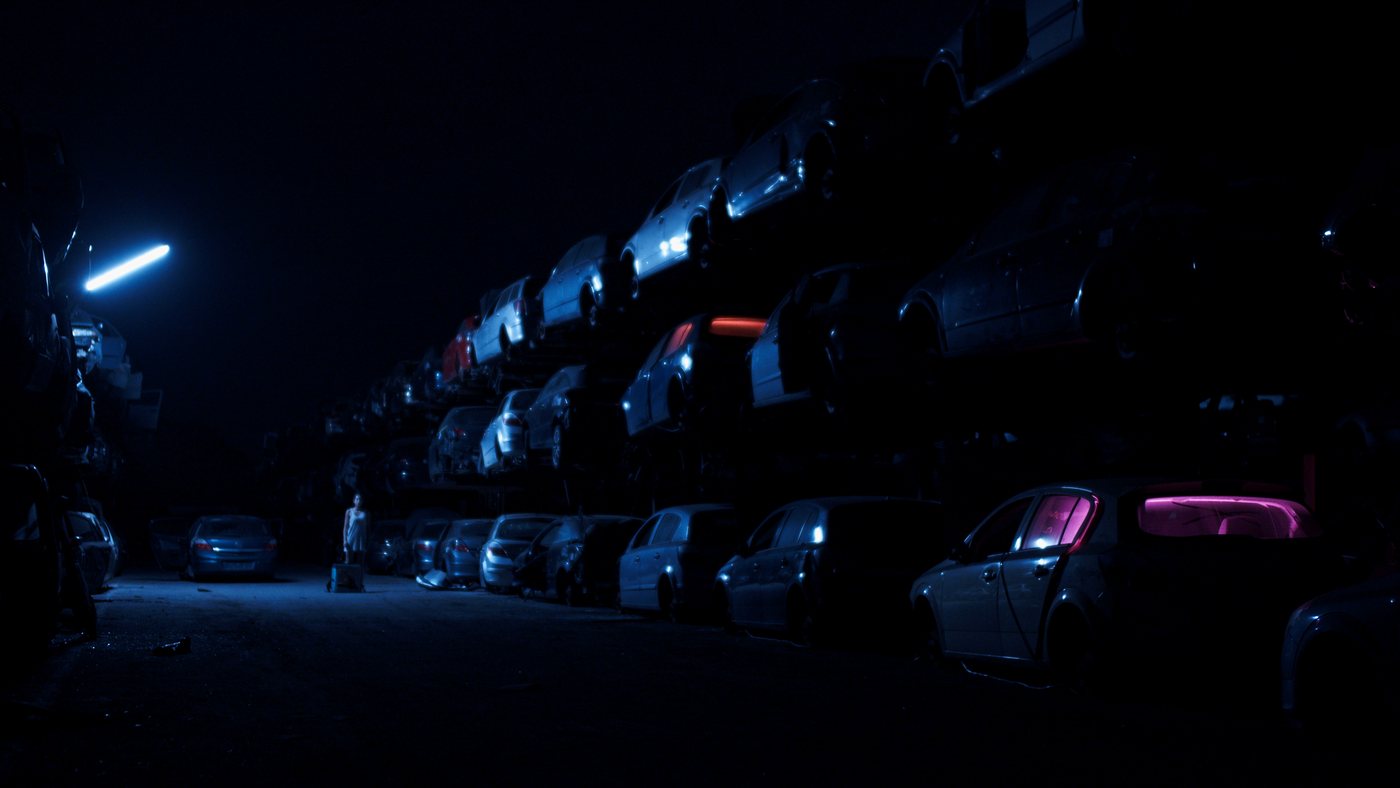 Ohnmacht — a lone figure among stacked cars under a fluorescent light