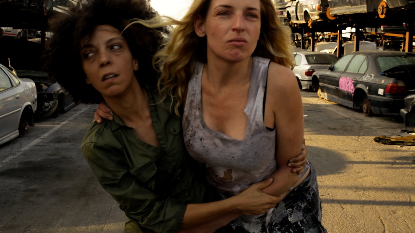Ohnmacht — a woman leads another through the scrapyard at sunset