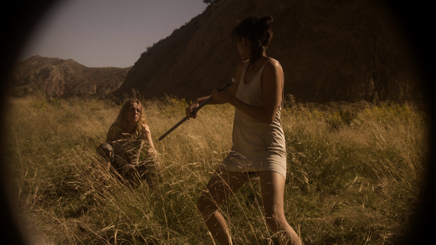 Ohnmacht — one woman holds a rifle over another kneeling in a dry grass field