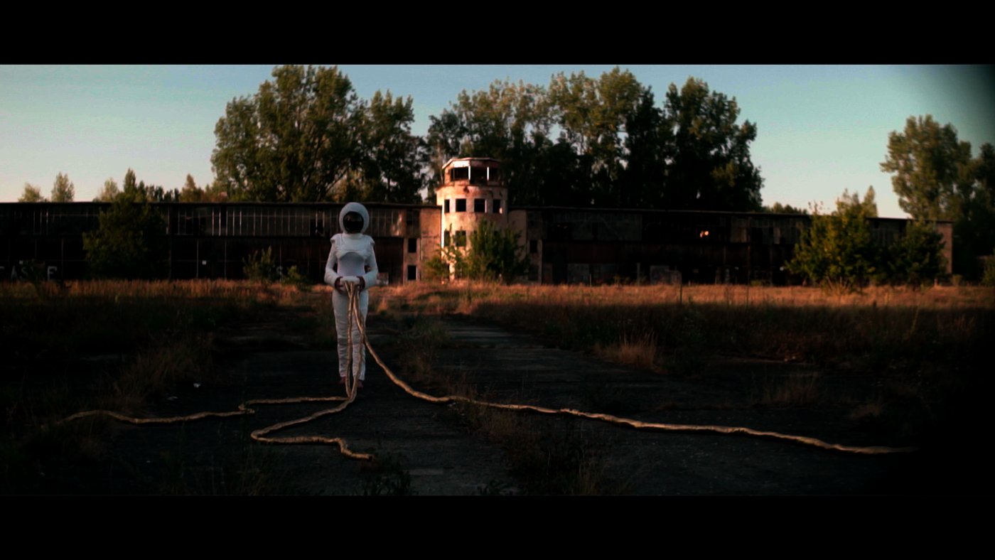 Ohnmacht — a figure in a white spacesuit stands before an abandoned building, a rope trailing across the ground
